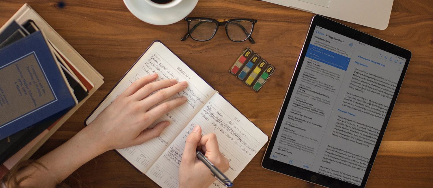 Picture of a woman journaling at a desk. To her left is a pile of books. To her right is an iPad . Around her there is also a cup of coffee, glasses, post-it flags, and a macbook.