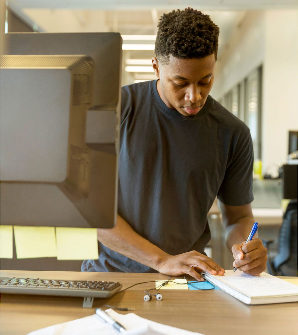 Young man writing on a notebook at a standing computer desk